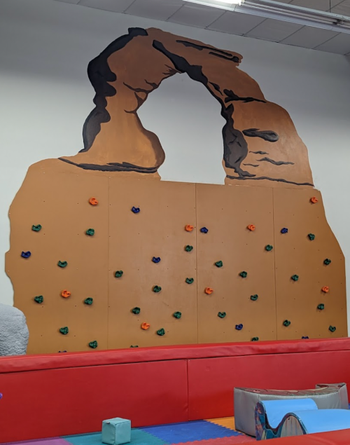 A colorful indoor climbing wall designed to resemble a rock formation, with soft padding below for safety.