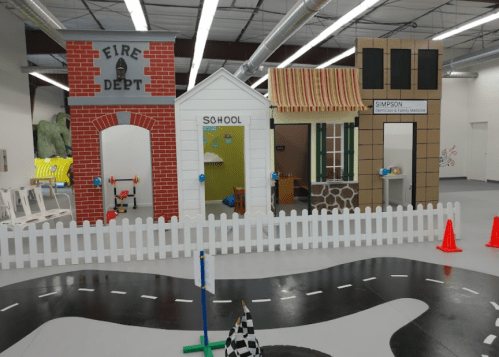 A colorful play area featuring a fire department, school, and store, with a toy road and safety cones.