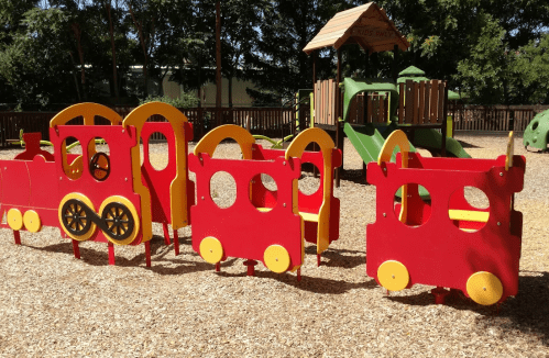 Colorful wooden train play structure in a playground, surrounded by wood chips and greenery.