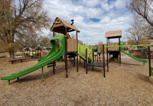 A playground with green slides and wooden structures, surrounded by gravel and trees under a cloudy sky.