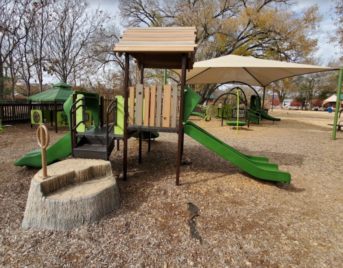 Playground with green slides, a wooden play structure, and a shaded area, surrounded by gravel and trees.