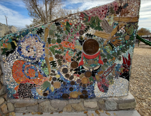 Colorful mosaic artwork featuring various patterns, coins, and nature elements, set against a stone wall.