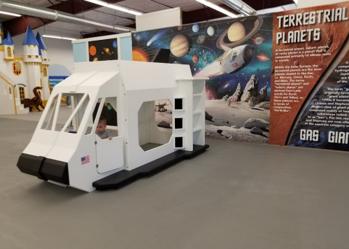 A child sits in a large, white cardboard spaceship in a colorful space-themed exhibit.