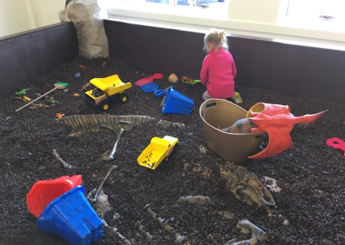 A child plays in a sandpit filled with toy trucks, colorful plastic items, and dinosaur bones.