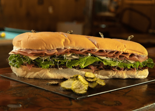A large sub sandwich filled with layers of deli meats, lettuce, and sprouts, served with pickles on the side.