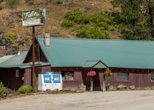 A rustic inn with a green roof and wooden exterior, featuring a sign and flower pots outside.
