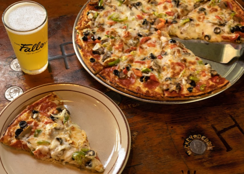 A pizza with various toppings on a plate, alongside a glass of beer on a wooden table.