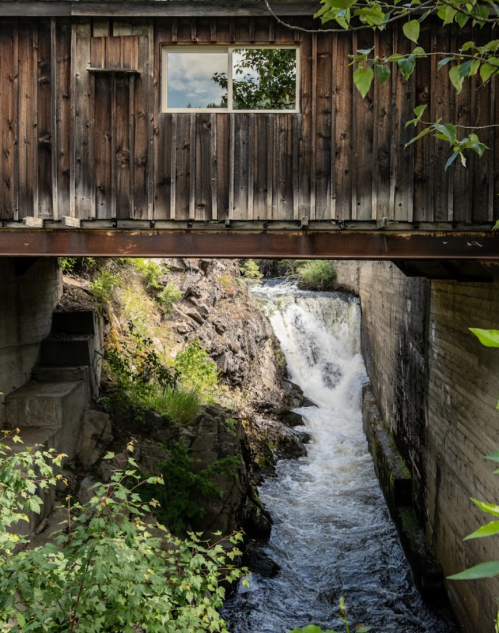 A wooden structure above a rushing waterfall, surrounded by greenery and rocky terrain.