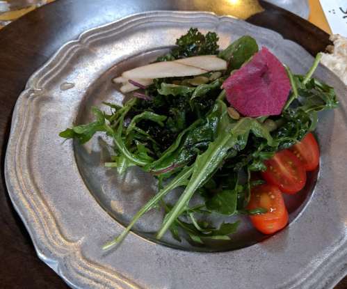A fresh salad with mixed greens, cherry tomatoes, sliced pear, and a pink flower garnish on a silver plate.