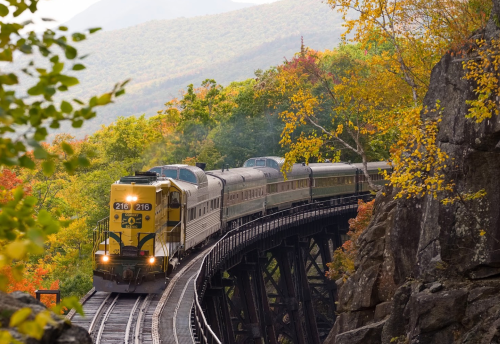 A train travels along a curved trestle bridge surrounded by vibrant autumn foliage and mountains in the background.