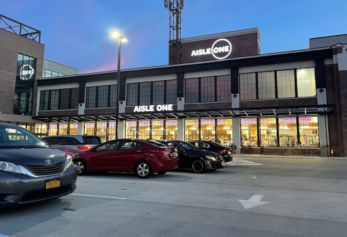 Exterior of a building labeled "AISLE ONE" at dusk, with parked cars in the foreground and a tower in the background.