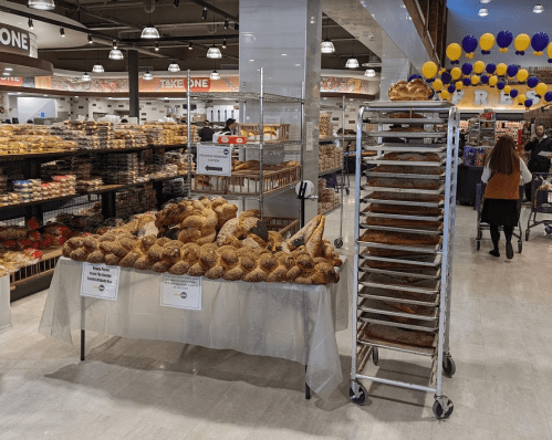 A bakery display with various breads on a table and a rack of baked goods in a bright store setting.