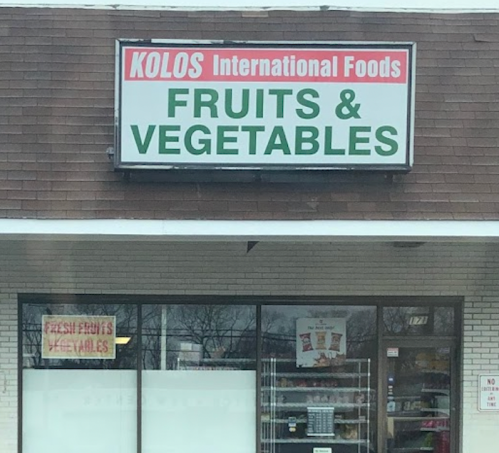 Sign for Kolos International Foods featuring "Fruits & Vegetables" with a window display of fresh produce.