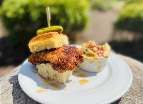 A plate with a biscuit sandwich featuring fried chicken and pickles, alongside a small cup of creamy dip.