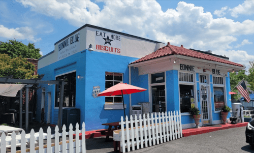 A bright blue restaurant with a red roof, white picket fence, and outdoor seating under umbrellas on a sunny day.