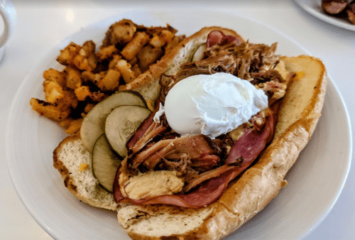 A sandwich with pulled pork, ham, and a poached egg, served with seasoned potato cubes and pickles on the side.