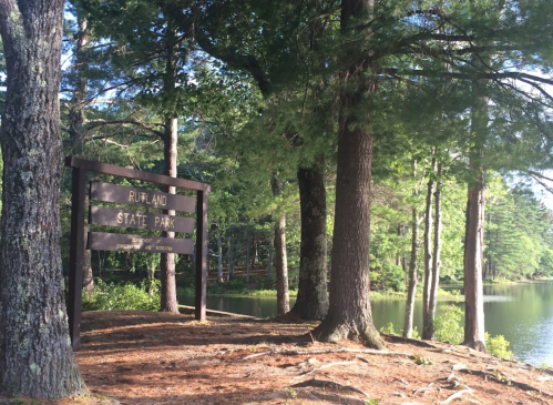 Sign for Rutland State Park surrounded by tall trees and a calm lake in the background.