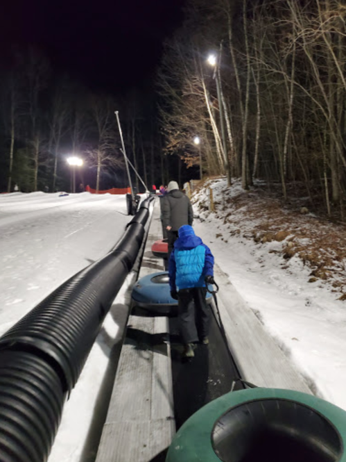 A child in a blue jacket walks up a snowy slope, pulling a snow tube, with trees and lights in the background.