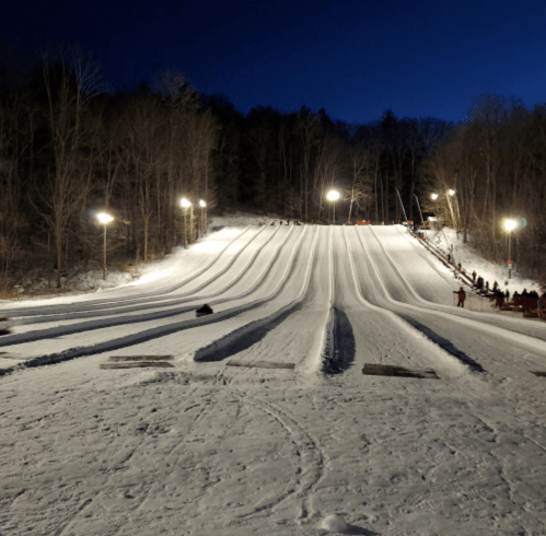 A snowy hill for tubing, illuminated by lights, with tracks leading down and people waiting in the background.