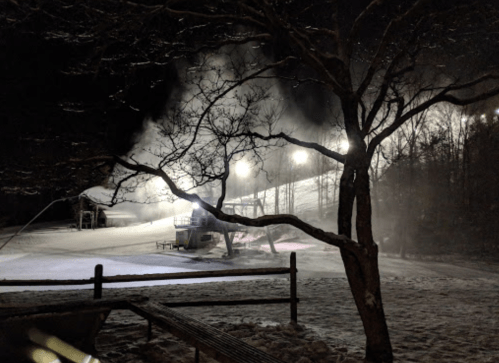 Snow-covered ski slope at night, with snowmaking machines and bright lights illuminating the scene.