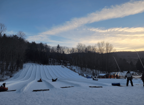 Snowy tubing hill with multiple lanes, trees in the background, and a sunset sky. People are preparing to slide down.