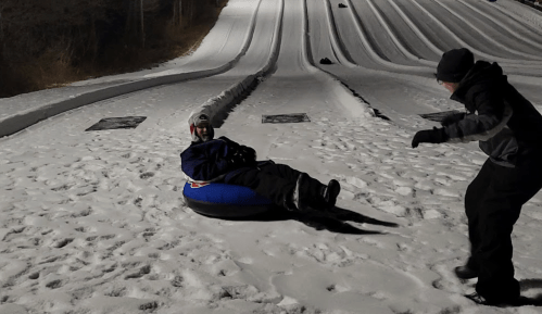 A person sits on a blue snow tube on a snowy slope, while another person playfully approaches from the side.