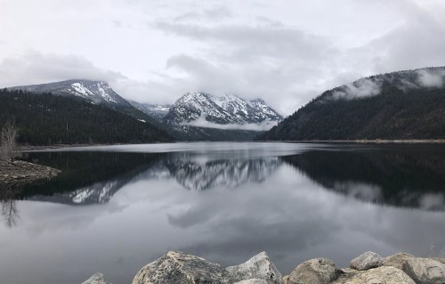 A serene lake reflects snow-capped mountains and cloudy skies, surrounded by lush green forests.