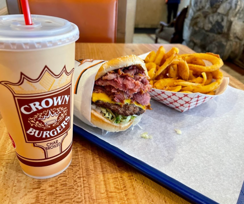 A burger wrapped in paper, topped with cheese and meat, served with curly fries and a drink on a tray.