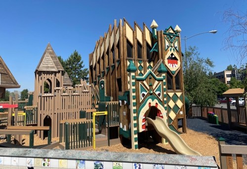 Colorful playground structure with slides and climbing areas, surrounded by wooden fencing and a clear blue sky.