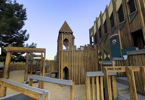 A wooden playground resembling a castle, featuring towers and slides, set against a clear blue sky.
