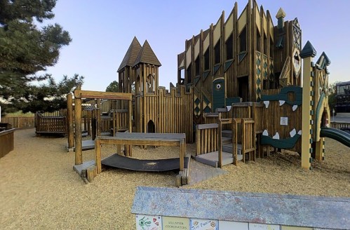 A wooden playground resembling a castle, featuring towers, slides, and climbing structures, set in a sandy area.