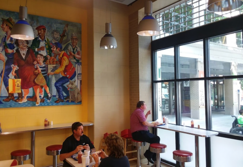A cozy diner with colorful wall art, featuring patrons enjoying meals at tables by large windows.