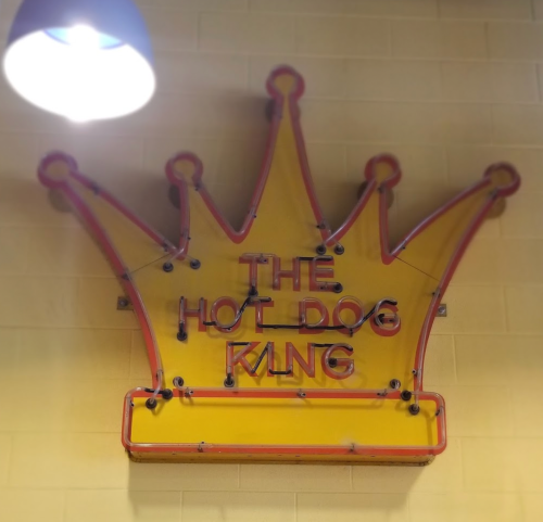 Neon sign of a crown with the text "THE HOT DOG KING" in bold letters, set against a yellow wall.