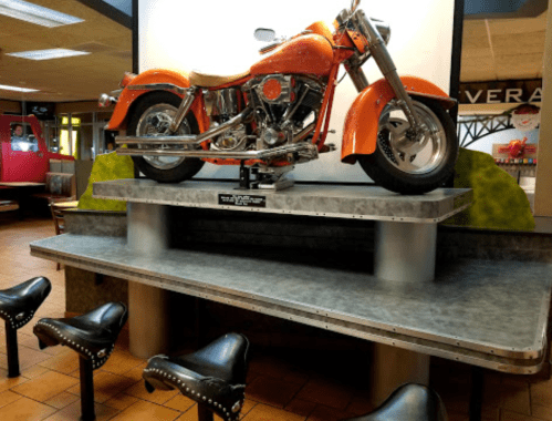 A vintage orange motorcycle displayed on a metal platform in a restaurant, with black stools in front.