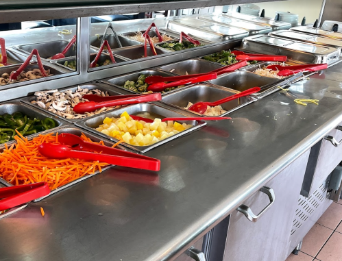 A salad bar with various fresh vegetables and fruits in metal trays, each with red serving utensils.
