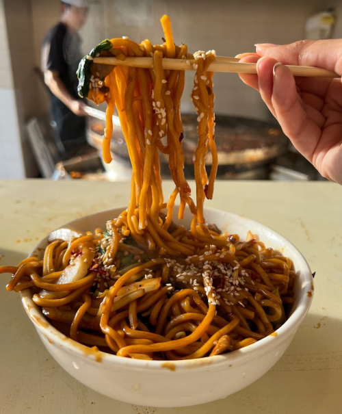 A hand holding chopsticks lifts noodles from a bowl, topped with sesame seeds and sauce, in a kitchen setting.