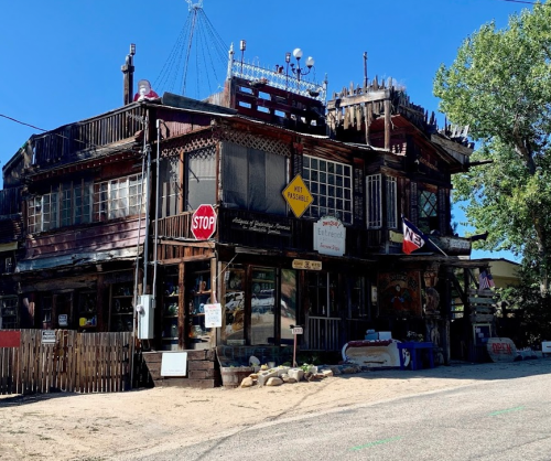 A rustic, multi-level building with wooden architecture, signs, and a sandy area in front under a clear blue sky.