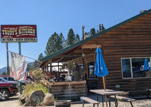 A rustic wooden building with a sign for Trudy's Kitchen, featuring outdoor seating and a scenic mountain backdrop.