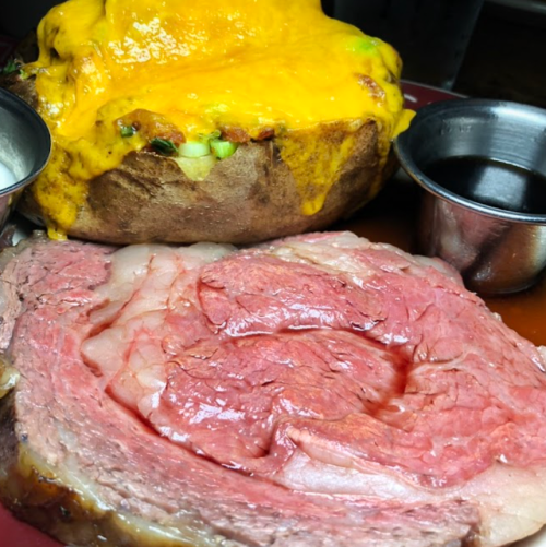A plate featuring a slice of pink prime rib next to a loaded baked potato topped with melted cheese.