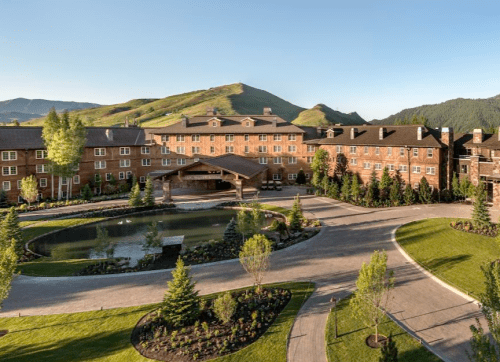 A scenic view of a brick hotel surrounded by greenery and a pond, with mountains in the background.