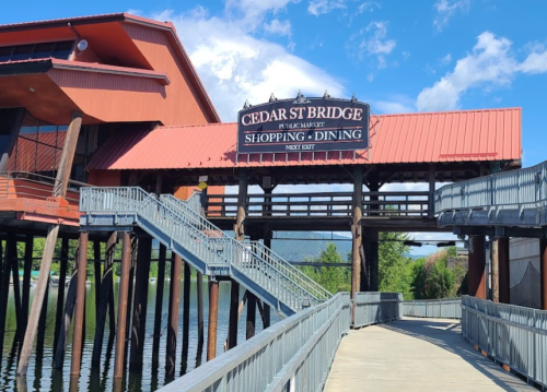 Cedar St Bridge sign above a wooden walkway, leading to shopping and dining, with water and trees in the background.