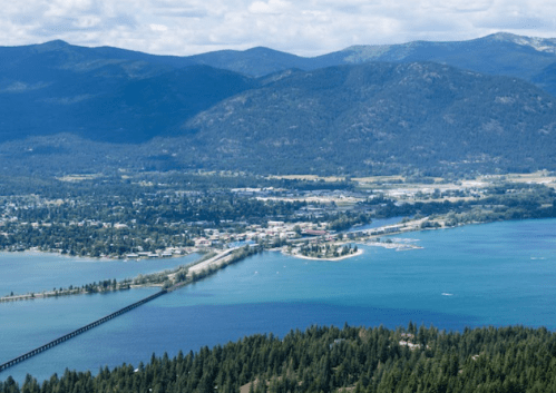 Aerial view of a town by a lake, surrounded by mountains and lush greenery, with a bridge connecting to the land.