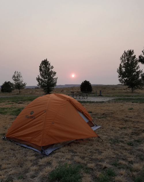 An orange tent sits on dry grass with a hazy sunset in the background, framed by distant trees.