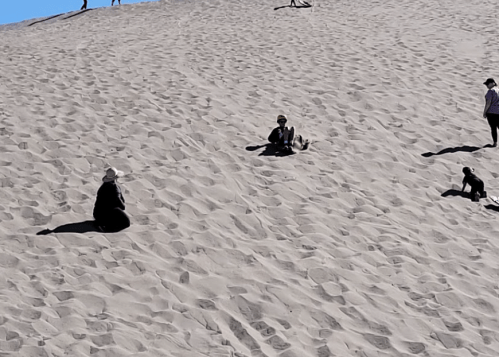 People sitting and playing on a sandy dune under a clear blue sky, with shadows cast on the sand.