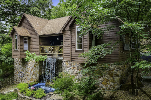 A charming wooden house on a stone foundation, featuring a waterfall and surrounded by lush greenery.