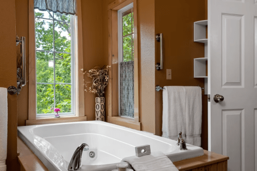 A cozy bathroom featuring a jetted tub, brown walls, a window with greenery outside, and neatly arranged towels.