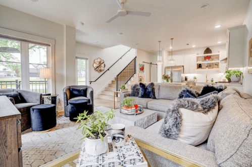 A modern living room with a gray sofa, black chairs, and a staircase leading to a bright kitchen. Plants and decor add warmth.