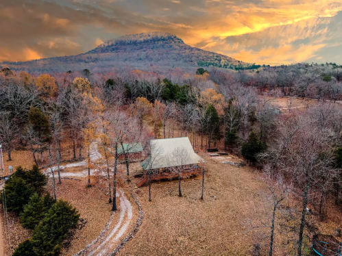 Aerial view of a rustic cabin surrounded by autumn trees, with a mountain in the background under a colorful sky.