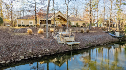 A lakeside home with a wooden deck, surrounded by trees and autumn foliage, reflecting in the calm water.