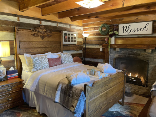 Cozy rustic bedroom with a wooden bed, warm lighting, and a stone fireplace, featuring decorative pillows and a "Blessed" sign.